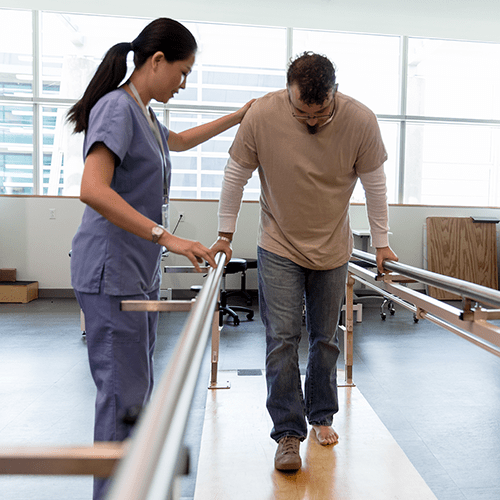 Adult male patient takes his first steps after foot surgery using the orthopedic parallel bars under the guidance of the female physical therapist.