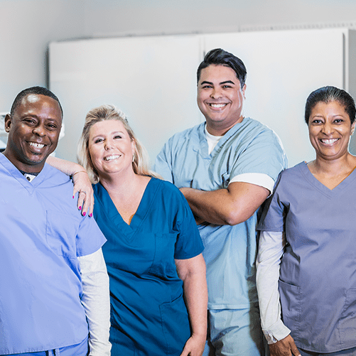 A group of four medical professionals working together. The team is standing side by side wearing scrubs, smiling at the camera.