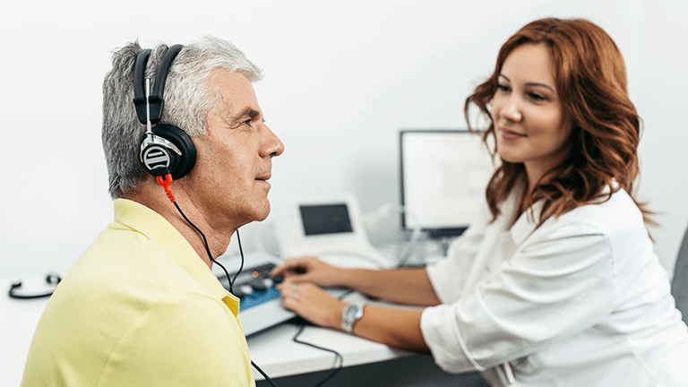 Senior patient wearing headphones talking to Audiologist during ear exam at doctors office.