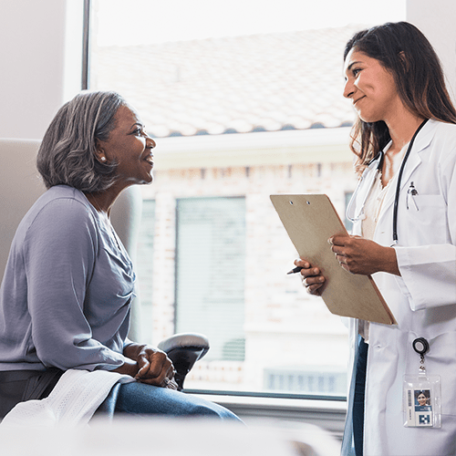 A caring female physician assistant smiles with compassion while talking with a senior female patient during a medical exam.