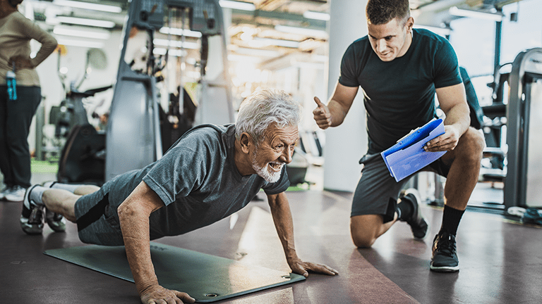 Man exercising push-ups in a gym while young fitness professional is training him.