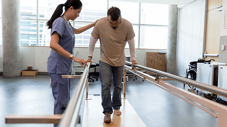 Adult male patient takes his first steps after foot surgery using the orthopedic parallel bars under the guidance of the female physical therapist.