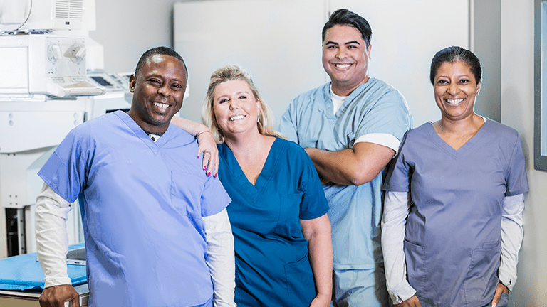 A group of four medical professionals working together. The team is standing side by side wearing scrubs, smiling at the camera.